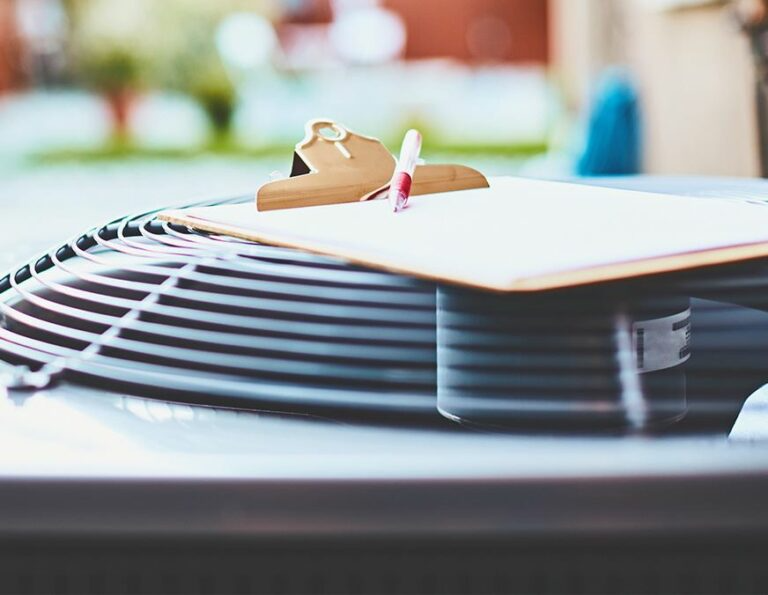This image shows a clipboard with a pen resting on top of the circular fan grille of an outdoor air conditioning unit.