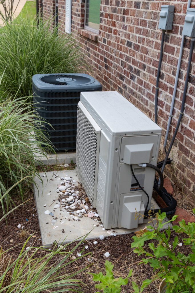 Two home AC units sit on a concrete pad near a brick house.