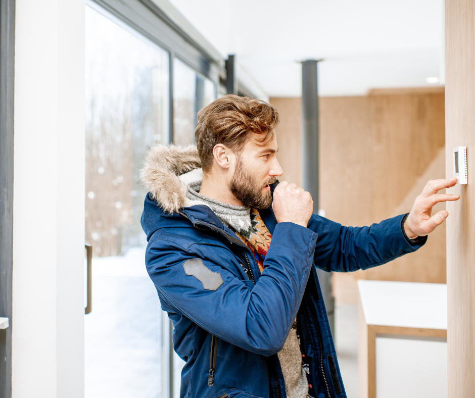 A man in a winter coat adjusts his thermostat while shivering, likely indicating a problem with his furnace.